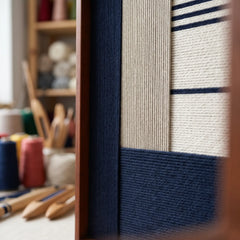 Angled close-up of the framed Baltic Window hand-woven tapestry showcasing the wooden frame and dimensional navy blue and beige wool patterns with yarn spools in the background.