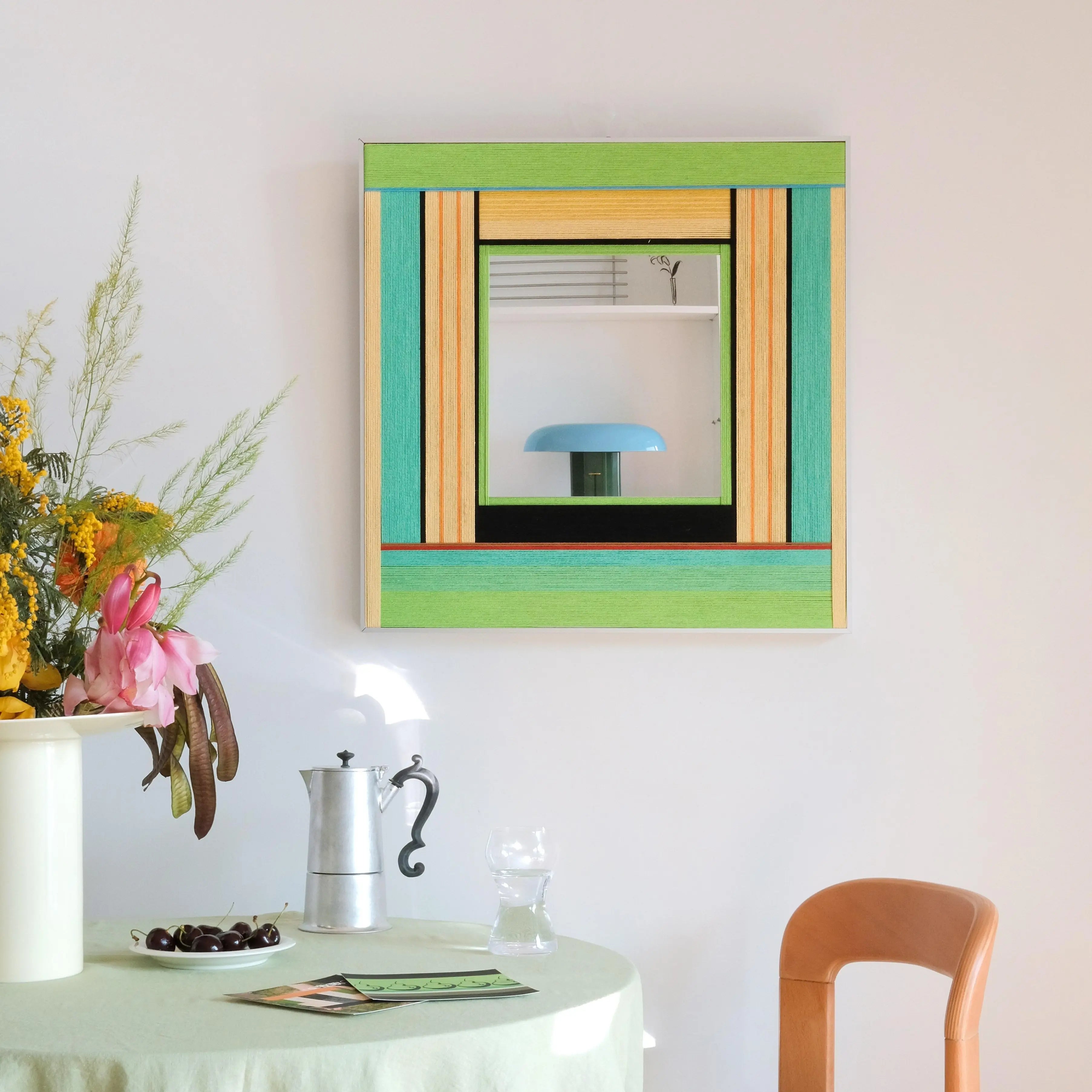 Breathing Like Fresh Grass hand‑woven yarn mirror wall art featuring alternating mint green, grass green, and natural wood stripe borders framing a central mirror panel, displayed above a small table with vase.