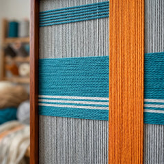 Macro close-up of the Breeze of Capri textile art highlighting the structural layering of the raised bright orange vertical yarn intersecting over a teal horizontal band.