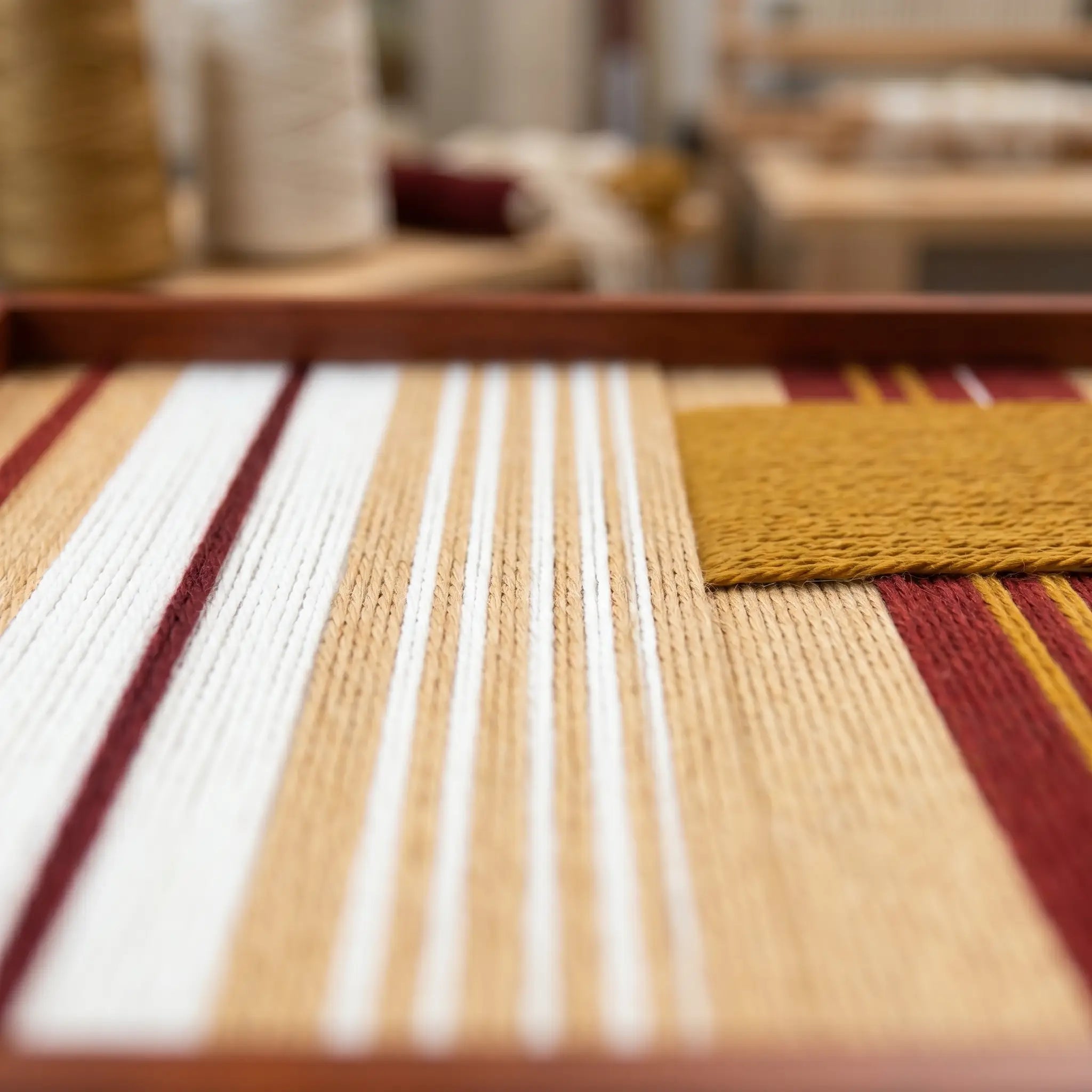 Extreme low-angle macro shot of the Carmine Pulse hand-woven textile art lying flat in a studio, highlighting the textured carmine red, mustard yellow, and white yarn stripes.