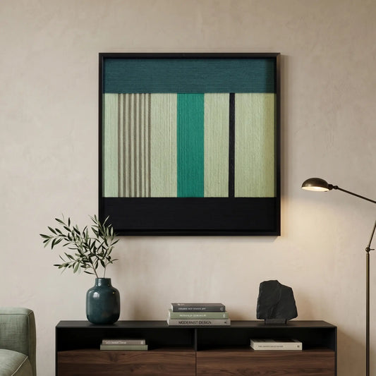 Square hand-woven wool wall art in a black frame featuring vertical stripes in forest green, sage, and emerald, hanging above a dark wood console with a black architect lamp.