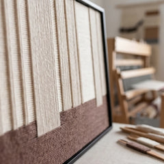 Angled close-up shot of the Ivory Rhythm textile art showcasing the raised dimensional cream yarn ending on a brown base, with weaving tools in the background.