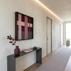 Framed textured fiber art displayed in a minimalist hallway above a sleek black console table with a burgundy vase.