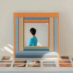 Square woven textile mirror reflecting a person in a blue shirt, styled resting on a table surrounded by scattered printed photographs in bright sunlight.