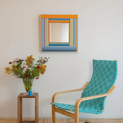 Miami Sunsets textile framed mirror hanging in a brightly lit room next to a mid-century teal armchair and a wooden stand with a colorful floral bouquet.