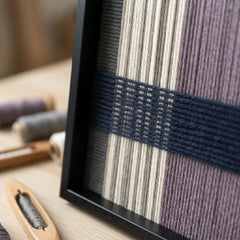 Angled close-up of the framed Midnight Lavender hand-woven tapestry showcasing the black frame and dimensional navy blue woven lines over cream and lavender wool.