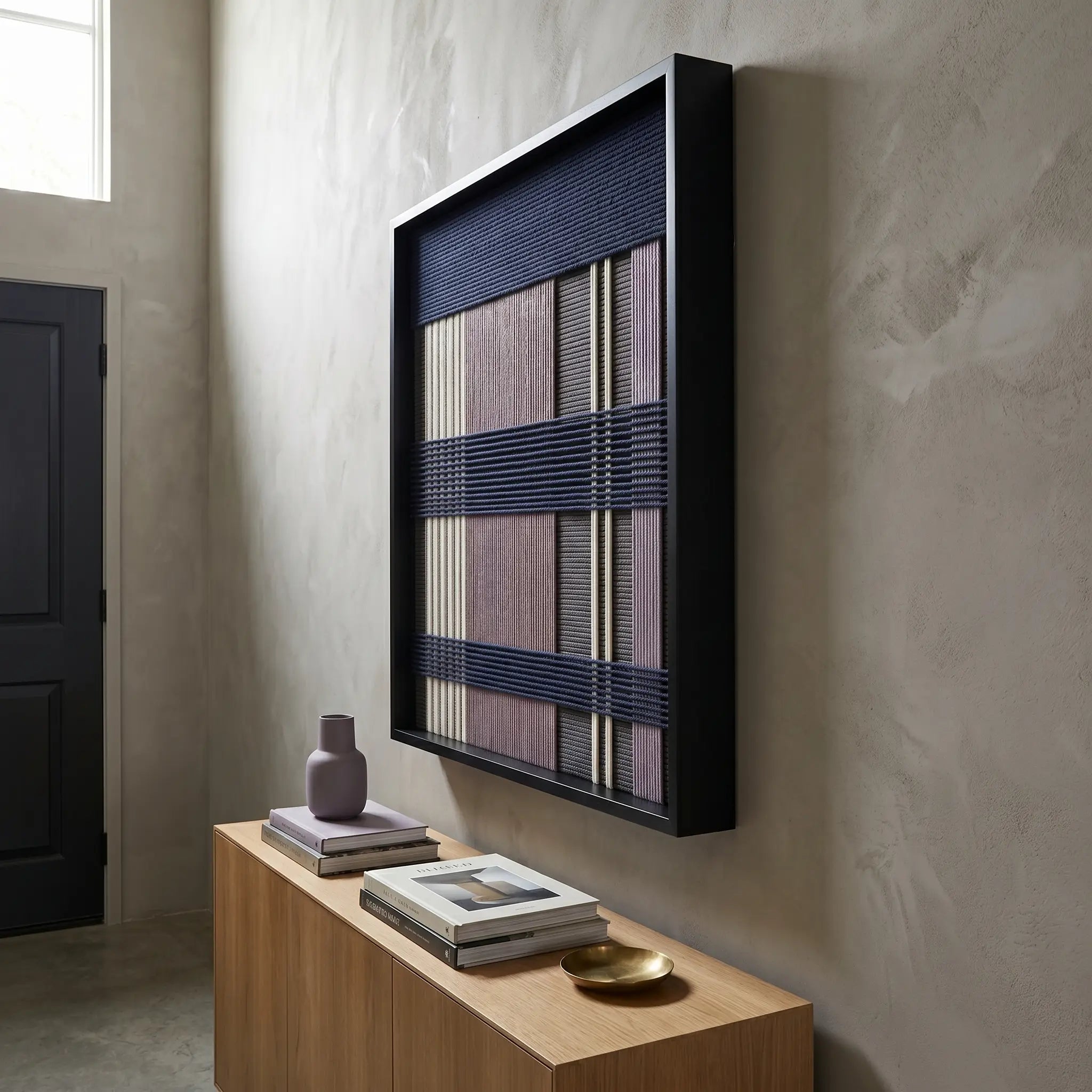 Angled view of the framed Midnight Lavender textile wall art hanging gracefully on a textured wall above a minimalist light wood console table.
