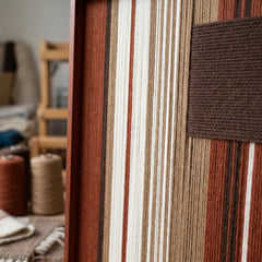 Angled close-up of the framed Mocha Prelude hand-woven tapestry showcasing the wooden frame and dimensional dark brown patch against striped earth-toned wool.