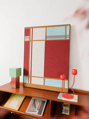 Red House yarn wall art styled on a desk beside stationery and a mint-green block ornament, showcasing its compact square format and vivid color blocking.