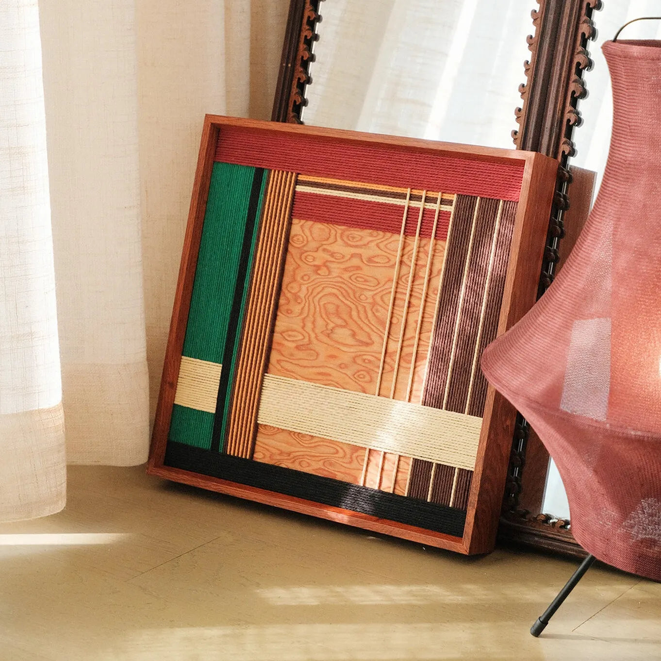 Saint Christmas hand-woven wool yarn wall art with alternating festive red, emerald green, and natural wood-tone stripes in a dark wood frame, displayed on a console table beside a violin and decorative plant