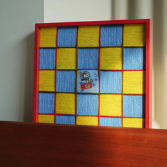 A focused shot of the red-framed wool tapestry resting on a dark wooden surface, highlighting the high contrast between the frame and woven texture.