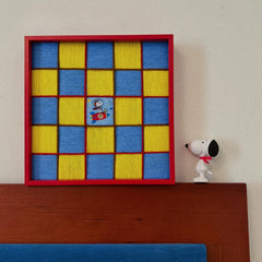 Red framed hand-woven wool wall art featuring a yellow and blue checkerboard pattern with a Snoopy rocket patch, displayed on a wooden headboard.