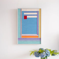 A portrait-oriented framed fiber art tapestry featuring colorful abstract wool weaving displayed on a clean white wall with flowers.