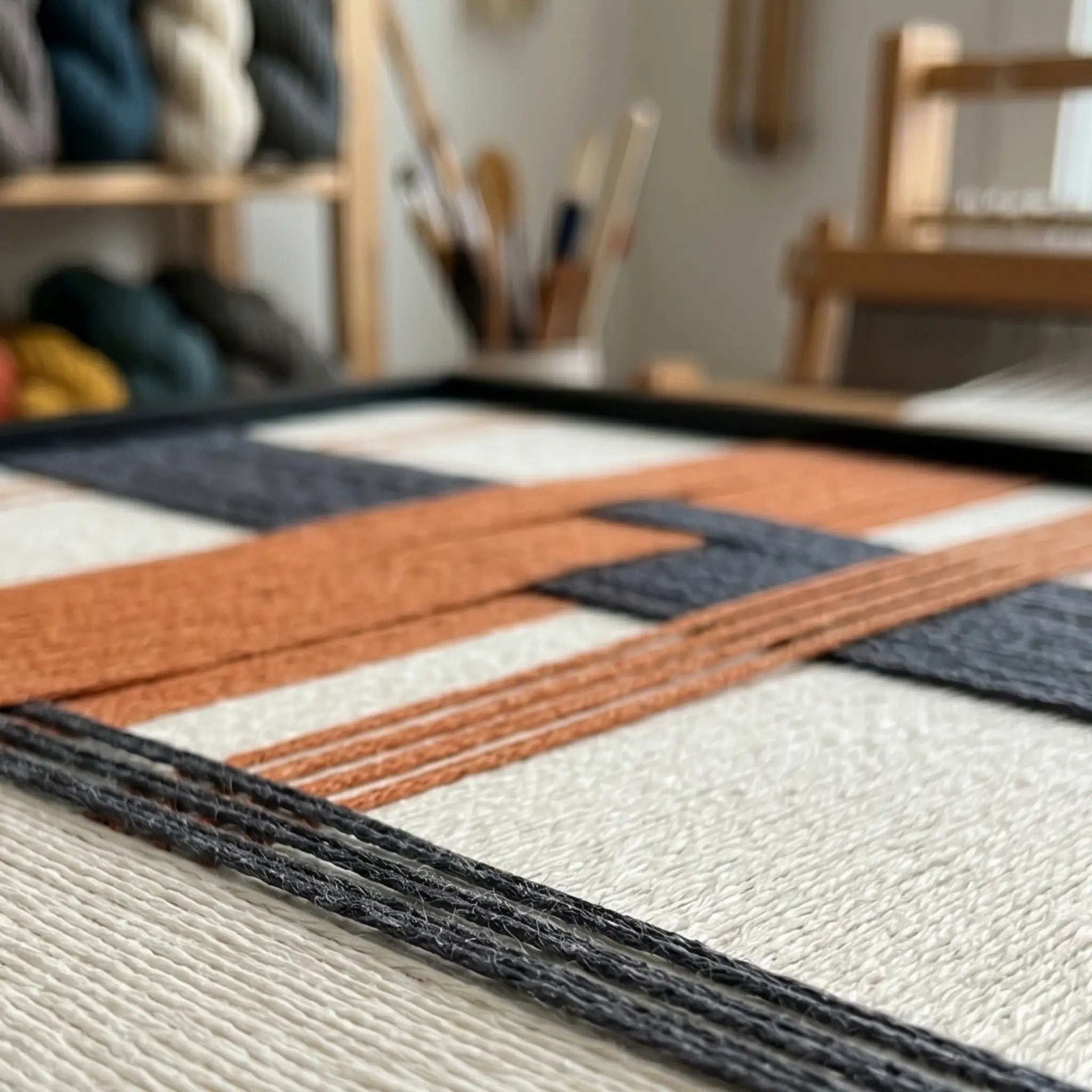 Macro close-up of the Terracotta Dusk fiber art in a workshop setting, highlighting the structural layering of the raised rust orange and charcoal yarn over the cream background.