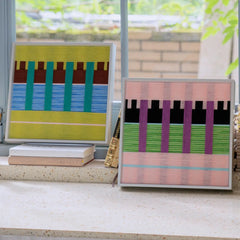 Green and blue woven textile art sitting on stacked books next to a pink variant on a low shelf near a bright window.