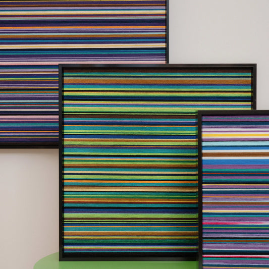 A diptych set of square hand-woven wool wall art featuring fine horizontal stripes in dark jewel tones, greens, and purples, styled in a moody modern living room.