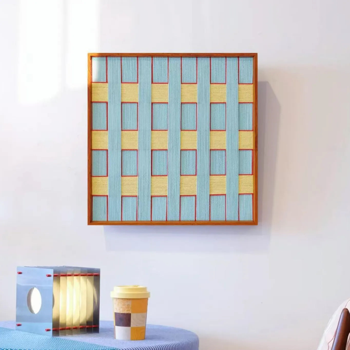 Framed light blue and yellow woven wall art hanging on a bright white wall above a small blue cafe table with a lamp and coffee cup.