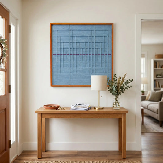 Framed blue textile wall art styled in a bright entryway above a light oak console table with a lamp and vase.
