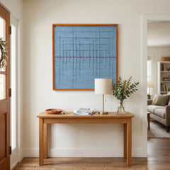 Framed blue textile wall art styled in a bright entryway above a light oak console table with a lamp and vase.