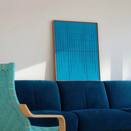 The textured blue fiber art piece displayed in a sunlit living room behind a dark blue velvet sofa, creating a calming monochromatic look.