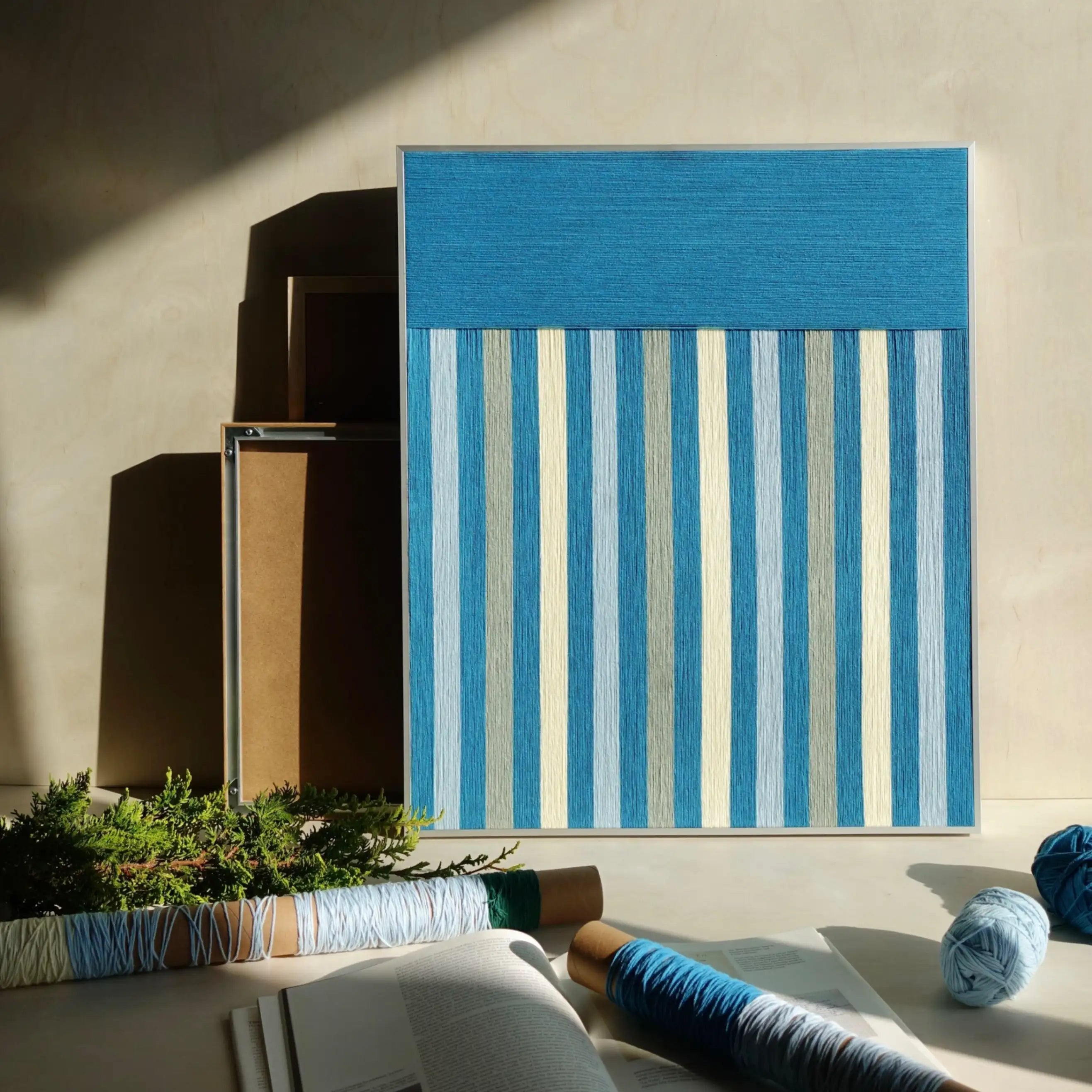 Blue Night hand-woven wool yarn wall art with alternating light and dark blue vertical stripes in a minimalist wood frame, displayed on a tabletop to showcase its precise geometric design
