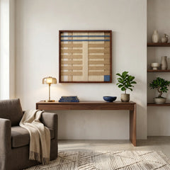 Sophisticated framed fiber wall art hanging above a minimalist wooden console table in a bright modern entryway with an armchair.