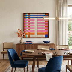 Colorful California Afterglow fiber wall art hanging above a wooden credenza in an open dining space featuring a round table and blue velvet chairs.