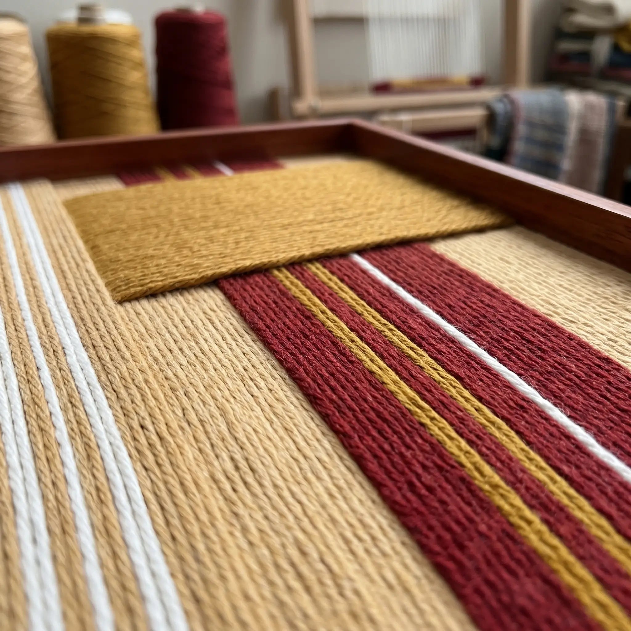Angled close-up shot of the Carmine Pulse textile art showcasing the raised dimensional mustard yellow patch over textured red and beige hand-woven wool stripes.