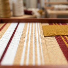 Extreme low-angle macro shot of the Carmine Pulse hand-woven textile art lying flat in a studio, highlighting the textured carmine red, mustard yellow, and white yarn stripes.
