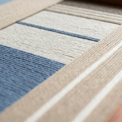 Extreme low-angle macro shot of the Cerulean Mist hand-woven textile art lying flat, emphasizing the raised blue, beige, and white striped yarn texture.