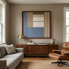 The Cerulean Mist hand-woven wool tapestry provides a striking focal point over a wooden console behind a grey sofa and a classic brown leather lounge chair.