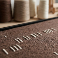 Angled close-up of the Cocoa Whispers textile art lying flat, showcasing the raised white yarn details against a brown woven canvas with yarn spools in the soft-focus background.