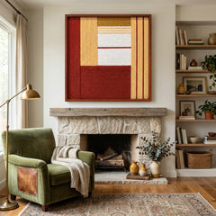 Framed Crimson Daybreak textile wall art displayed in a cozy living room, hanging above a rustic stone fireplace mantel next to a classic green armchair.