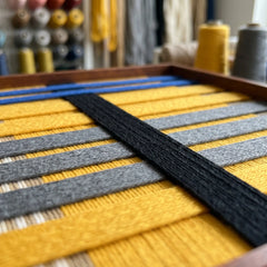 Angled close-up of the Golden Monsoon tapestry in a workshop setting, showcasing the dimensional depth of the hand-wrapped black, yellow, and blue yarn.