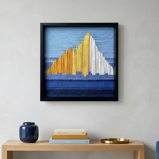 Square hand-woven wool wall art in a black frame depicting an abstract mountain landscape with golden and white peaks against a blue sky, hanging above a light wood console table.