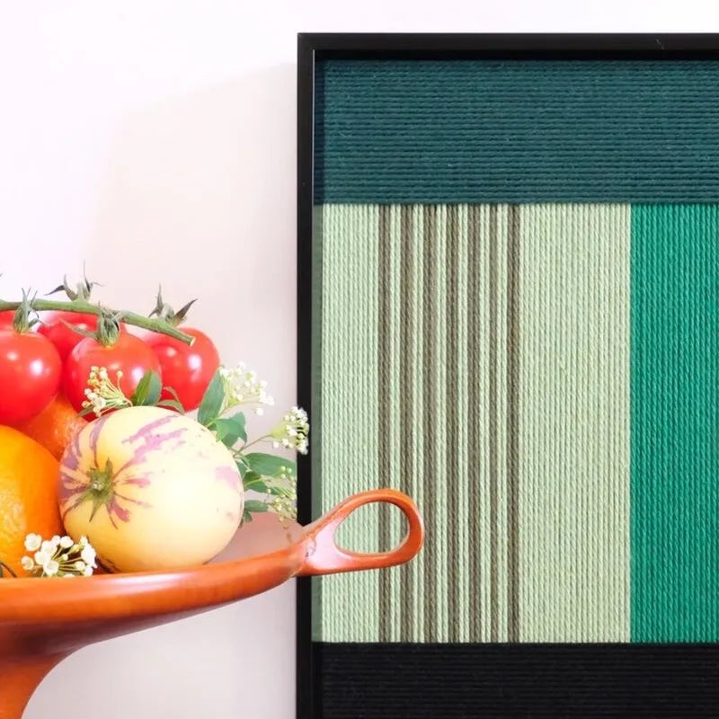 Green Branches yarn wall art styled on a desk next to a fruit bowl and black sculpture, emphasizing vivid color contrast and tactile woven texture.
