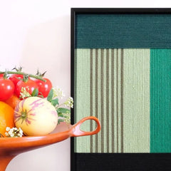 Green Branches yarn wall art styled on a desk next to a fruit bowl and black sculpture, emphasizing vivid color contrast and tactile woven texture.

