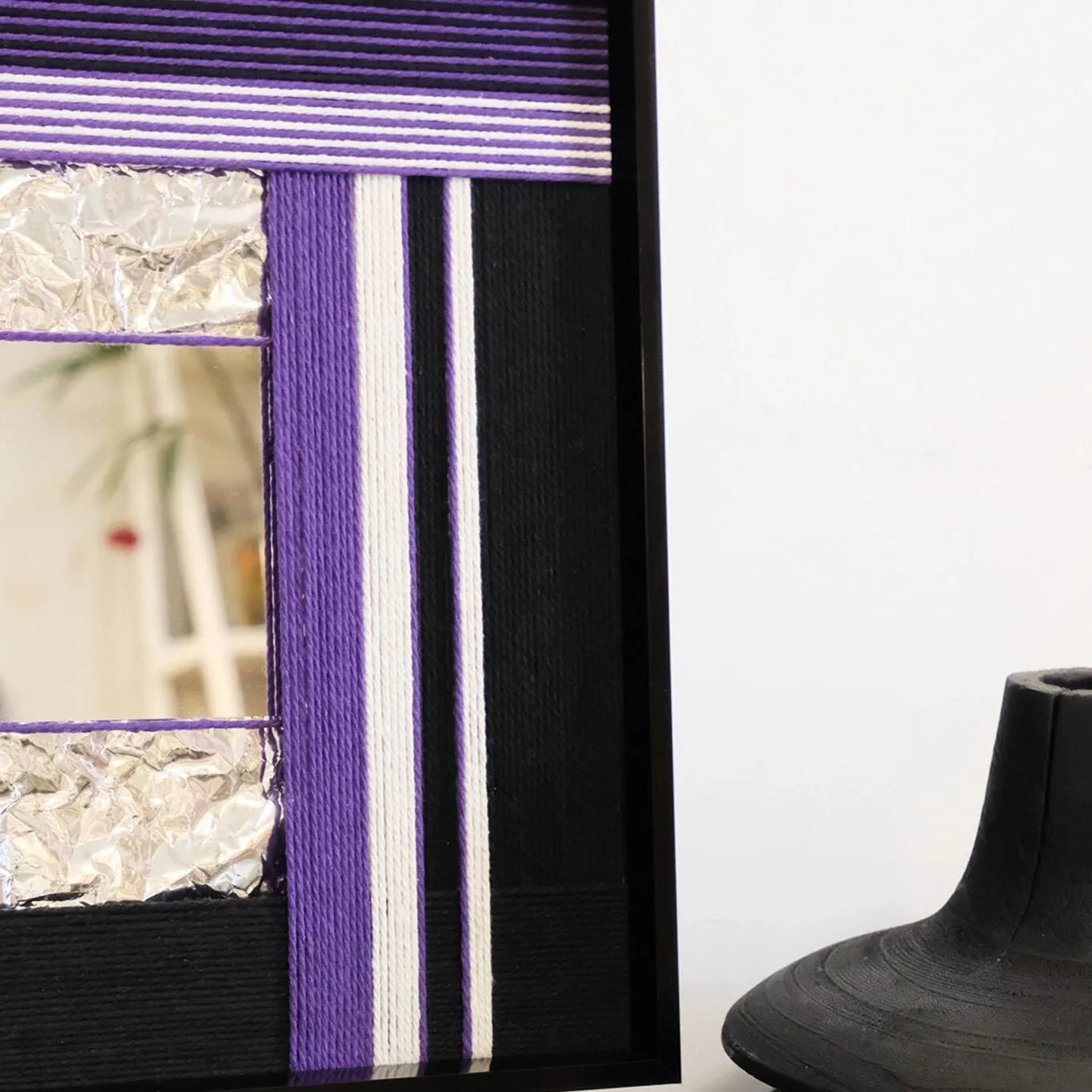 Angled view of HOUTIAN yarn mirror art leaning on a tabletop, highlighting the contrast between purple yarn stripes and black frame with integrated mirror.
