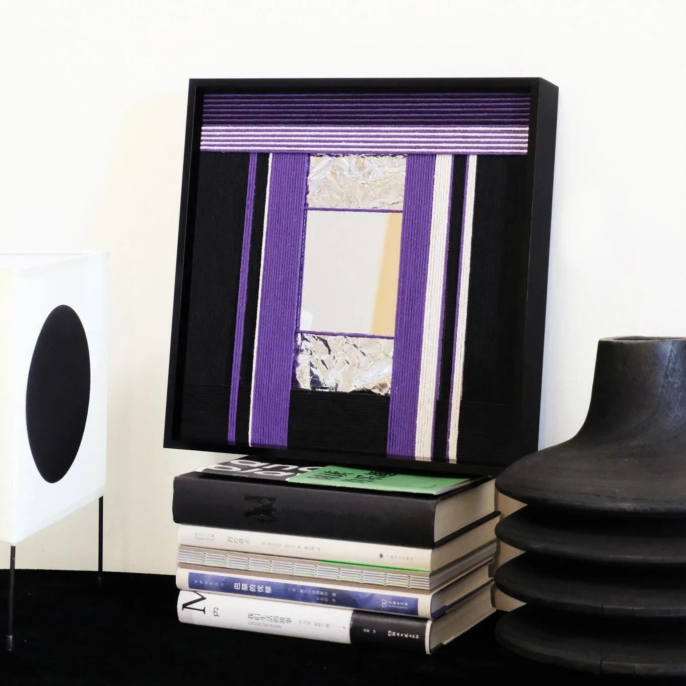 HOUTIAN hand-woven wool yarn mirror wall art with alternating purple and black vertical stripes framing a central mirror panel, displayed on a console table between speaker and books.
