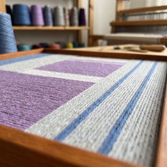 Hand-woven wool tapestry displayed in an artist studio with spools of blue and grey yarn and a weaving loom in background.