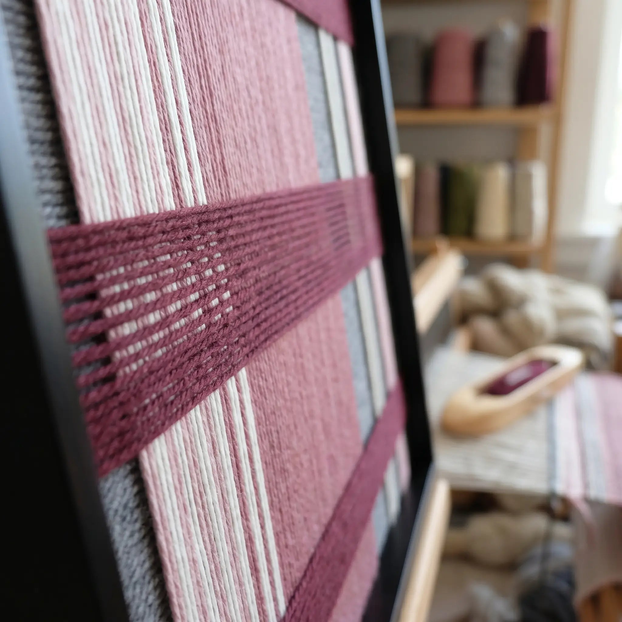 Angled close-up of the framed Merlot Veil hand-woven tapestry showcasing the black frame and dimensional deep red horizontal bands over pink and grey vertical wool stripes.