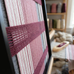 Angled close-up of the framed Merlot Veil hand-woven tapestry showcasing the black frame and dimensional deep red horizontal bands over pink and grey vertical wool stripes.