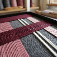 Extreme angled macro close-up of the Merlot Veil hand-woven textile art lying flat, highlighting the textured burgundy, pink, and grey yarn over white warp threads.