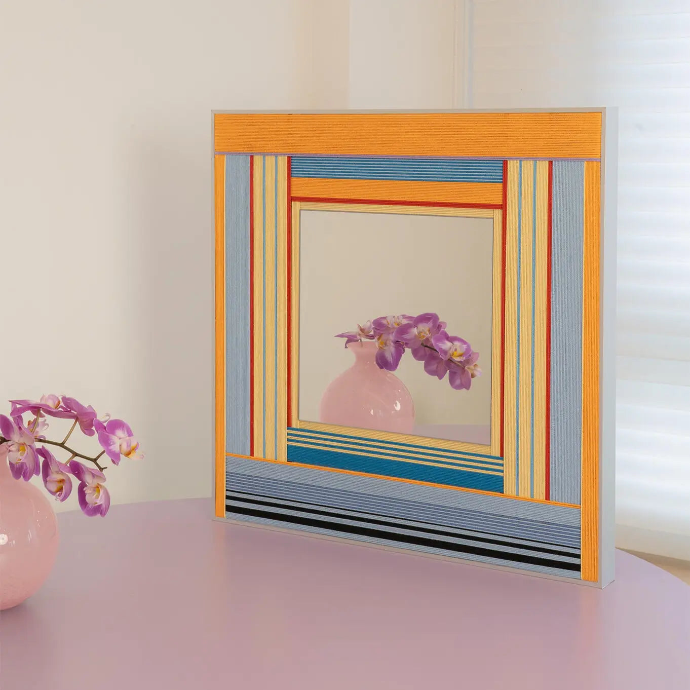Angled perspective of the Miami Sunsets textile mirror resting on a pastel purple table, reflecting a beautiful pink orchid plant.