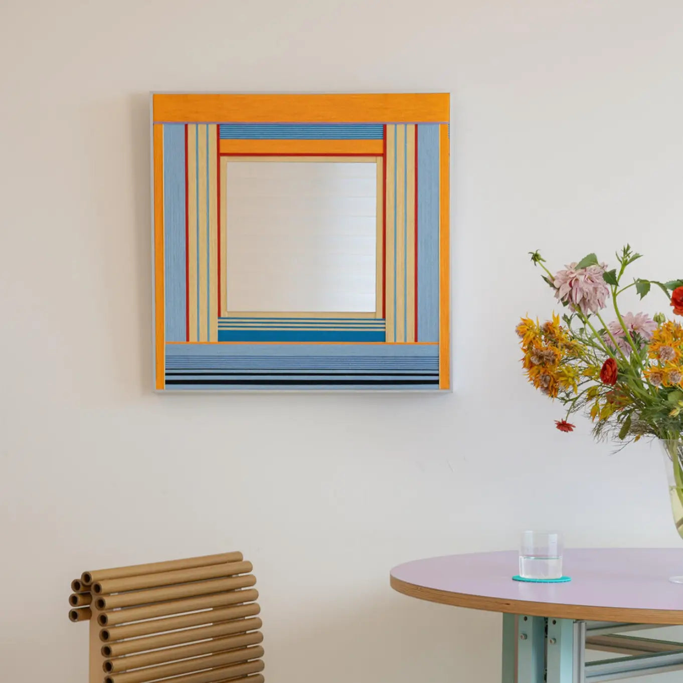 Square Miami Sunsets textile mirror art featuring a woven border with vibrant orange, blue, and pastel stripes hanging above a round purple table with a floral arrangement.