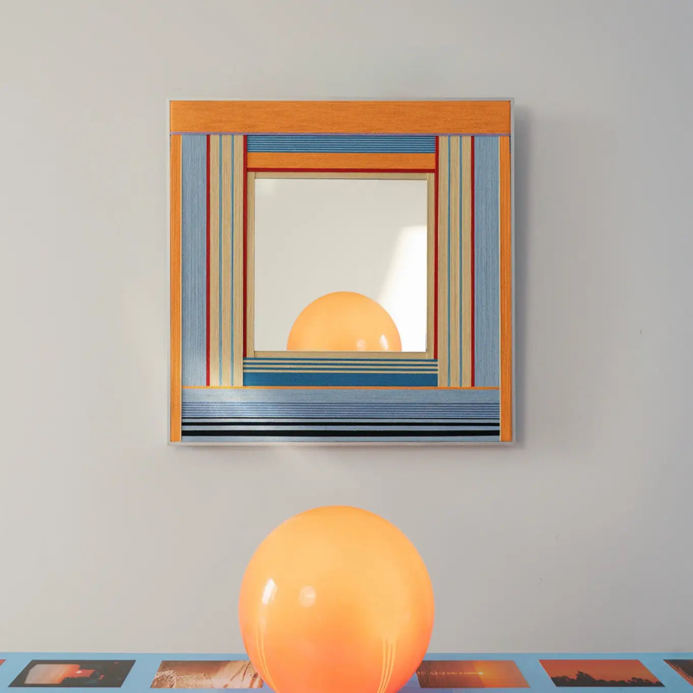 Framed textile mirror art with an orange and blue woven border reflecting a bright orange balloon resting on a blue surface.