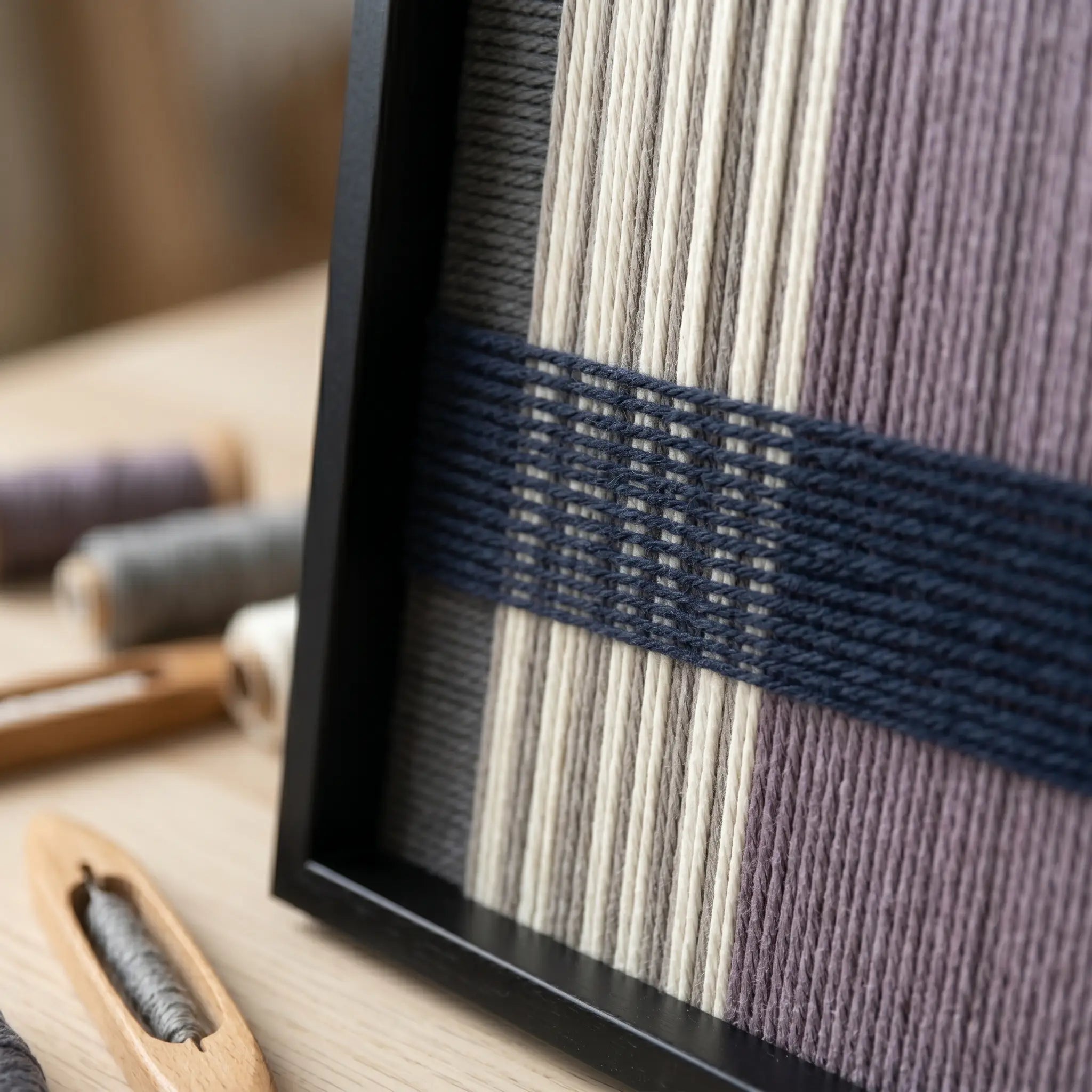 Angled close-up of the framed Midnight Lavender hand-woven tapestry showcasing the black frame and dimensional navy blue woven lines over cream and lavender wool.