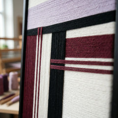 An angled close-up view of the framed fiber art in a studio, showcasing the dimensional layering of the lavender and maroon wool.