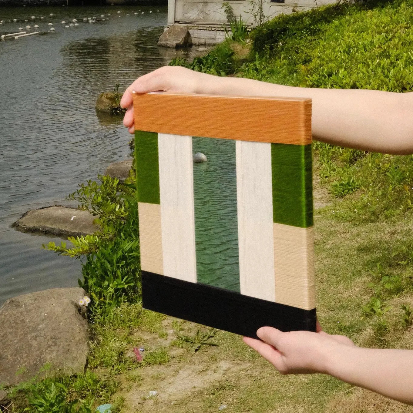 Pastoral Elegance yarn wall art held by water’s edge, emphasizing the natural wood frame and contrasting green-beige panel design
