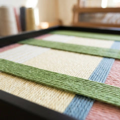 Low-angle close-up of the framed fiber wall art in a studio setting, showcasing the dimensional depth and structural multi-directional layering of the yarn.